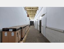 Langer, schmaler Lagerflur mit weiß gestrichenen Wänden, einer offenen Holzbalkendecke und Betonboden. Auf der linken Seite stehen Kartons auf Paletten gestapelt, auf der rechten Seite führen Türen zu angrenzenden Räumen.