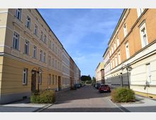 Eine ruhige Straße mit gepflastertem Gehweg, gesäumt von Reihen historischer Wohnhäuser in gelben und roten Fassaden. Auf beiden Seiten stehen Laternen und parkende Autos, im Hintergrund sind Bäume und blauer Himmel sichtbar.