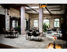 Geräumiger Innenraum eines Restaurants oder Cafés im Industriestil mit unverputzten Ziegelwänden, großen Fenstern, freiliegenden Deckenbalken und modernen Hängelampen. Der Raum ist eingerichtet mit Holztischen, schwarzen Stühlen und dekorativen Pflanzen.