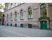 Ziegelsteinfassade eines historischen Gebäudes mit mehreren großen, grün umrandeten Rundbogenfenstern sowie einer grünen Doppeltür. Der Bürgersteig davor ist von herabgefallenem Herbstlaub bedeckt, und links ist ein Torbogen zu sehen.