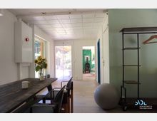 Innenraum eines hellen Wohnbereichs mit Holztisch und Stühlen, einem großen Fenster im Hintergrund, das auf eine Terrasse führt. Im angrenzenden Raum sind eine grüne Wand mit Blättermuster und ein Kaminofen sichtbar, daneben ein Regal und ein Gymnastikball.