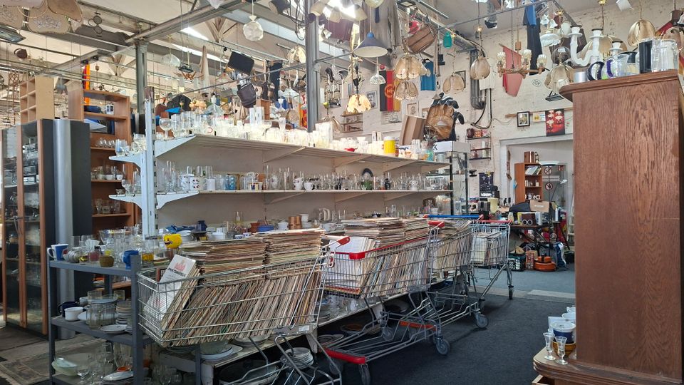 Innenansicht eines vollgestellten Trödelladens in einer hohen, industriellen Halle mit Metallstützen und vielen von der Decke hängenden Lampen und Leuchten. Regale und Tische sind dicht mit Gläsern, Geschirr und Kleinteilen gefüllt; davor stehen Einkaufswagen mit gestapelten Schallplatten.