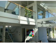 Modernes, mehrstöckiges Wohninnere mit viel Tageslicht, weißen Wänden und großen Glasflächen. Zu sehen sind eine offene Treppe, ein Balkon mit Glasgeländer, eine Zimmerpflanze und ein farbenfrohes abstraktes Gemälde an der Wand.