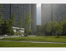 Grüne Parkfläche mit jungen Bäumen im Vordergrund, umgeben von mehrspurigen Straßen und parkenden Autos; im Hintergrund zwei hohe Wohnhochhäuser in moderner Architektur sowie ein niedrigeres Gebäude mit flachem Dach.