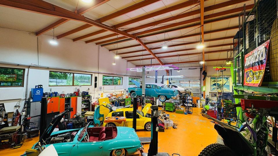Große Autowerkstatt mit hohen Deckenbalken, orangefarbenem Boden und vielen Werkbänken, Regalen sowie Hebebühnen. Mehrere Oldtimer und Fahrzeuge stehen im Innenraum, teils mit geöffneten Motorhauben, neben Werkzeug und Ersatzteilen.