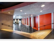 Offener Eingangsbereich mit moderner, minimalistischer Architektur, gekennzeichnet durch glatte Betonwände, orangefarbene Akzente und eine breite Treppe. Eine runde Deckenöffnung ermöglicht Lichteinfall, und im Hintergrund befindet sich ein gläserner Eingang.