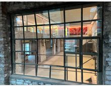 Ansicht eines Industriegebäudes mit Blick durch ein großes, gitterförmiges Fenster auf einen offenen Innenraum mit freiliegenden Backsteinwänden, Betonboden und einer Zwischendecke. Im oberen Bereich befinden sich farbige Lichtinstallationen und Wandkunst, unten sind offene Treppen und freie Flächen sichtbar.