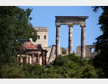 Ruine eines historischen Gebäudes inmitten von Bäumen, mit markanten Säulen im vorderen Bereich, einer Backsteinstruktur und einer teilweise erhaltenen steinernen Fassade im Hintergrund. Der blaue Himmel kontrastiert mit der grünen Vegetation und den alten Mauern.