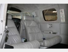 Innenraum eines kleinen Flugzeugs mit grauen Ledersitzen, einer Konsole an der Wand mit Lüftung und Armlehne sowie einem Fenster. Im Hintergrund ist das Cockpit mit Steuerinstrumenten sichtbar.