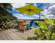 Holzterrasse am Wasser mit einem runden Tisch, umgeben von bunten Gartenstühlen und einem großen, grünen Sonnenschirm. Im Hintergrund sind ein See, bewaldete Ufer und ein Steg mit Booten zu sehen, während Topfpflanzen den Vordergrund schmücken.