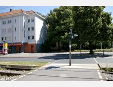 Wohngebäude mit hellem Putz und rotem Ziegeldach an einer Straßenkreuzung, daneben eine Ampel für Fußgänger mit grünem Signal. Im Vordergrund verlaufen Straßenbahnschienen, rechts befinden sich große, grüne Bäume.