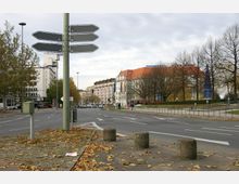 Straßenszene in einer Stadt mit einer breiten, geteerten Fahrbahn und Gehwegen, gesäumt von Bäumen ohne Laub. Im Hintergrund sind mehrstöckige Gebäude zu sehen, darunter ein Hotel mit rotem Dach und Flaggen davor, sowie ein blauer Turm als auffällige architektonische Struktur.