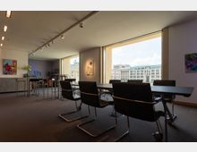 Innenraum eines modernen Konferenzraums mit schwarzen Stühlen und einem großen Tisch in der Mitte. Große Fenster bieten einen Blick auf das Brandenburger Tor und umliegende Gebäude.