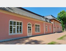 Ein lang gestrecktes Gebäude mit rosa Fassade, großen weißen Sprossenfenstern und einem Dach mit grauen Ziegeln. Der Vorplatz ist mit orangefarbenen Pflastersteinen bedeckt, und im Hintergrund sind grüne Bäume zu sehen.