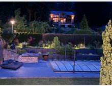 Abendaufnahme eines Gartens mit beleuchteter Poolabdeckung im Vordergrund und einer Sitzfläche aus Stein mit Pflanzenbeeten drumherum. Im Hintergrund steht ein modernes, zweistöckiges, beleuchtetes Holzhaus, umgeben von Bäumen und Hecken.