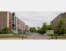 Straße mit Wohnblöcken auf beiden Seiten; links ein Gebäude mit farbigen Balkonen, rechts ein schlichtes Wohnhaus. In der Mitte eine asphaltierte Straße, gesäumt von parkenden Autos und Bäumen. Ein Container mit Graffiti und eine Baustellenabsperrung sind rechts im Bild zu sehen.