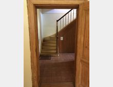 Holztreppenhaus mit sichtbar abgenutzten Stufen, aufgenommen durch einen rustikalen Türrahmen aus Holz. Der Boden besteht aus rötlichen Fliesen, und die Wandgestaltung ist schlicht, mit einer dezenten Bordüre im oberen Bereich.