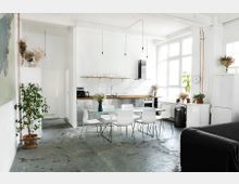 Heller, offener Wohnraum mit Küchenbereich, ausgestattet mit weißen Schränken, Holztresen und Glasregalen. Im Vordergrund ein Esstisch mit weißen Stühlen, Pflanzen in Töpfen und große Fenster, die viel Tageslicht hereinlassen.