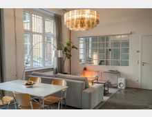 Wohnzimmer in einem modernen Loft-Stil mit industriellen Elementen. Zu sehen sind ein Esstisch mit Holzstühlen, ein graues Sofa, große Fenster mit weißen Rahmen, eine Hängelampe aus Glas, eine Zimmerpflanze und eine Wand mit großen Fensterpaneelen, die als Raumteiler dienen.
