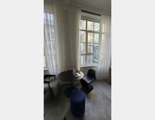 Heller Innenraum mit großen Sprossenfenstern und weißen, halbtransparenten Vorhängen an einer freigelegten Ziegelwand. Vor den Fenstern steht ein runder Tisch mit Stühlen und Hocker; durch die Fenster sind ein Innenhof mit Backsteinfassaden und eine Außentreppe zu sehen.