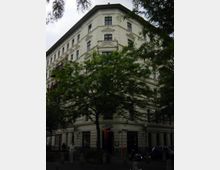 Eckgebäude im klassischen Stil mit heller, reich verzierter Fassade, mehreren Fenstern und einem Geschäft im Erdgeschoss. Vor dem Gebäude stehen hohe Bäume, die teilweise die Ansicht verdecken, sowie geparkte Autos und Personen.