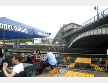 Ein Außenbereich eines Cafés oder Biergartens mit Holztischen und Stühlen, teilweise unter blauen Sonnenschirmen. Im Hintergrund sind ein Fluss mit einer Eisenbahnbrücke sowie ein großes Gebäude mit einer Glasfassade zu sehen.