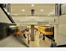 U-Bahn-Station mit einem gelben Zug der Linie U6, der in Richtung Alt-Mariendorf fährt. Menschen laufen auf dem Bahnsteig, die Wände sind mit weißen Fliesen verkleidet, und ein Schild weist die Fahrtrichtung Alt-Tegel aus.
