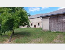 Ein ländlicher Hof mit einer grauen, einfachen Fassade und zwei kleinen Fenstern mit gemauerten Fensterrahmen; daneben ein Holzscheunentor. Im Vordergrund steht ein Baum mit grünen Blättern auf einer Wiese, der Himmel ist teils bewölkt.
