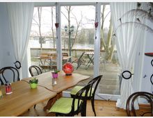 Esszimmer mit einem Holztisch und Stühlen, die grüne Sitzpolster haben, vor bodentiefen Fenstern, die auf eine Terrasse mit Gartenmöbeln und Blick auf einen Fluss führen. Im Hintergrund sind kahle Bäume und eine Brücke sichtbar.