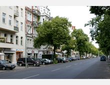 Straßenansicht mit mehrstöckigen Wohngebäuden im klassischen Stil, teils mit Balkonen, und einer Reihe von kleinen Geschäften im Erdgeschoss. Auf beiden Seiten der Straße stehen Bäume, unter denen Autos geparkt sind.