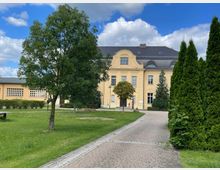 Gelbes historisches Gebäude mit großen Fenstern und einem geschwungenen Giebel, umgeben von grünen Bäumen und gepflegter Rasenfläche. Ein gepflasterter Weg führt durch den parkähnlichen Garten.