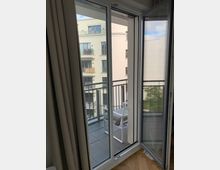 Eine geöffnete Balkontür in einem modernen Wohnraum, die zu einem kleinen Balkon mit Fliesenboden führt. Im Hintergrund sind mehrere beige Wohngebäude mit Balkonen und Fenstern zu sehen.