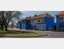 Straßenszene mit blauen Wohngebäuden und roten Ziegeldächern, umgeben von einer gepflasterten Straße und einer kleinen Grünfläche mit Bäumen. Die Gebäude haben weiße Fensterrahmen und einfache Fassadengestaltung.