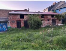 Verwilderter Hof mit hohem Gras vor einem niedrigen Backstein-Nebengebäude mit Wellblechdach, mehreren dunklen Holz­türen und kleinen Fensteröffnungen. Links steht ein blaues Garagentor mit Graffiti, rechts sind angrenzende Wohnhäuser und eine freistehende Badewanne im Außenbereich zu sehen.