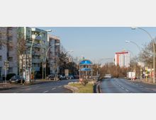 Straßenszene in einer städtischen Umgebung mit mittelhohen Wohngebäuden auf der linken Seite, einer zentralen U-Bahnstation mit dem Schild „U Rudow“ und einer zweispurigen Straße mit Straßenlaternen und Verkehrsampeln. Im Hintergrund sind moderne Hochhäuser und Bäume zu erkennen.