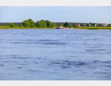 Weitläufige Flusslandschaft mit Bäumen und Wiesen am Ufer, im Hintergrund kleine Häuser und ein Frachtschiff auf dem Wasser unter klarem Himmel.