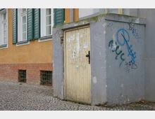 Ein kleiner grauer Anbau mit einer beigen Holztür, die mit Graffiti und Aufklebern bedeckt ist, steht vor einer Hauswand mit gelber Putzfassade, grünen Fensterläden und roten Ziegelsteinen an der Basis. Der Boden besteht aus Kopfsteinpflaster.