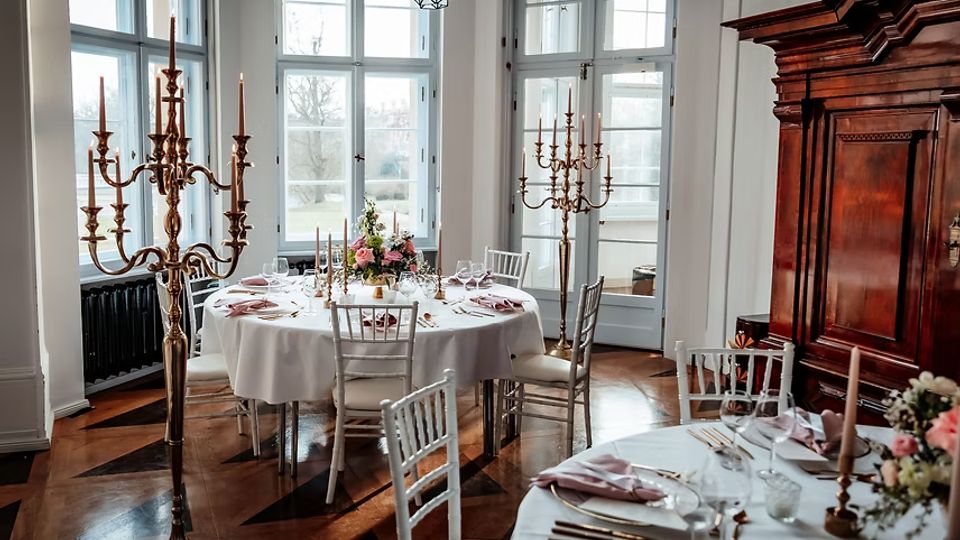Eleganter Speisesaal mit hohen, weiß gerahmten Fenstern und Holzboden, darin runde, weiß gedeckte Tische mit Blumenarrangements, Gläsern und Besteck. Mehrarmige goldene Kerzenständer und ein großer dunkler Holzschrank prägen den Raum.