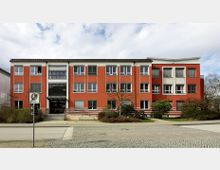 Rathausgebäude in moderner Architektur mit roter Fassade und großen weißen Fenstern, umgeben von kleinen Büschen und Bäumen; im Vordergrund führt ein Gehweg mit Straßenlaterne zum Eingang.