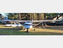 Ein kleines einmotoriges Flugzeug mit Schwimmern steht auf einer Freifläche in einem Waldgebiet. Im Hintergrund sind ein weiteres Flugzeug, ein militärisches Fahrzeug und Bäume erkennbar.