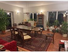 Wohnzimmer mit einem großen Holztisch und mehreren Stühlen auf einem orientalischen Teppich in der Mitte des Raumes. Die Wände sind beige, dekoriert mit Pflanzen, einem Glasschrank, einer Stehlampe und großen Fenstern, die teilweise mit Vorhängen verdeckt sind; zusätzlich hängt eine Gitarre an der Wand.