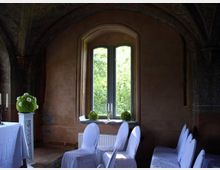 Ein historisch wirkender Raum mit einem großen Fenster, durch das grünes Laub sichtbar ist. Stühle mit weißen Hussen und dekorative Elemente wie Blumenarrangements und Kerzenständer befinden sich im Raum, dessen Wände und Decke mit dezenten Verzierungen gestaltet sind.