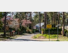 Straßenszene in einem Wohngebiet mit Einfamilienhäusern, umgeben von Bäumen und blühenden Sträuchern. Im Vordergrund stehen ein Ortsschild mit der Aufschrift „Wildau“ und ein Verkehrsschild für eine 30er-Zone, daneben Straßenschilder mit den Namen „Fontaneallee“ und „Kochstraße“.