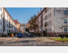 Wohnstraße mit mehrstöckigen Wohnhäusern aus heller Putz- und Ziegelsteinfassade auf beiden Seiten, geparkten Autos entlang der Straße und vereinzelten Bäumen. Straßenschilder und ein Fußgängerüberweg sind im Vordergrund zu sehen.