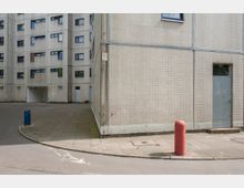 Außenecke eines grauen Wohnblocks mit kleinen quadratischen Fliesen, mehreren blauen Fenstern und einer Stahltür. Im Vordergrund sind ein Gehweg, ein roter Poller und eine gebogene Straßenkante zu sehen.