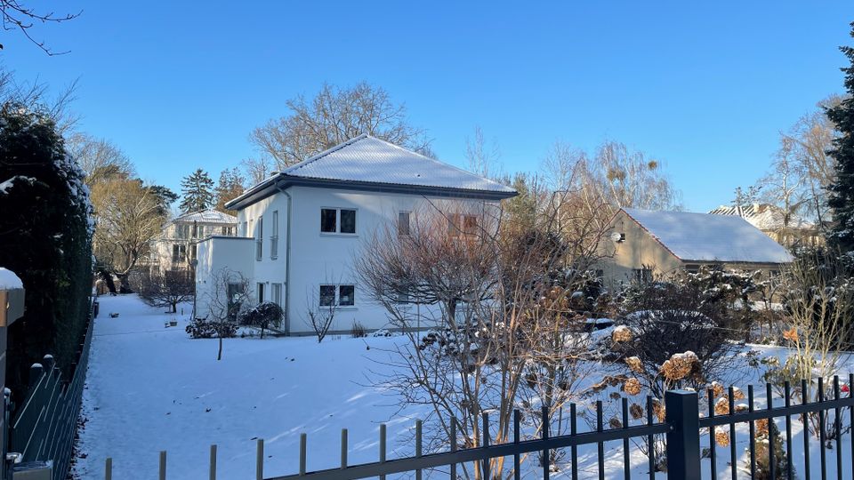 Schneebedeckter Garten mit Sträuchern und kahlen Bäumen vor einem weißen Wohnhaus mit Walmdach und mehreren Fenstern. Im Vordergrund verläuft ein Metallzaun, im Hintergrund sind weitere Häuser und Bäume unter klarem blauem Himmel zu sehen.