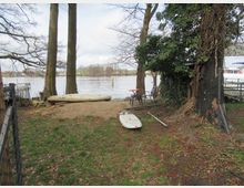 Grüne Uferlandschaft mit Bäumen, die einen sandigen Bereich und eine Wiese säumen. Im Vordergrund liegen ein Kajak und ein Surfbrett, während im Hintergrund ein ruhiger Fluss und ein Steg mit einem Boot sichtbar sind.