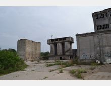 Verlassener Industriebereich mit mehreren verwitterten Betongebäuden, darunter ein Turm mit großen zylindrischen Stützen. Die Umgebung ist überwuchert von Gras und Sträuchern, und einige Wände sind mit Graffiti bedeckt.