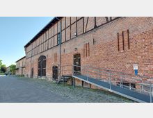 Historisches Backsteingebäude mit Fachwerkelementen und dunklen Holztüren, davor eine Rampe und Treppe aus Metall. Der Bereich ist gepflastert, und im Hintergrund sind Bäume sichtbar.