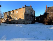 Zweistöckiges, beiges Gebäude mit Putzfassade und mehreren Fenstern, flankiert von einem seitlich angebauten Treppenaufgang. Im Vordergrund eine verschneite Wiese, daneben ein angrenzendes Gebäude mit teilweise bewachsener Fassade.